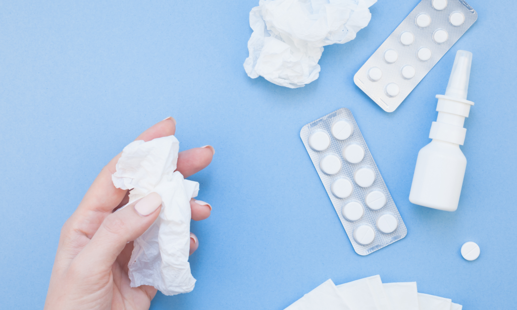 flatlay of hand holding tissue on a blue background, with other tissues, tablets and nasal spray